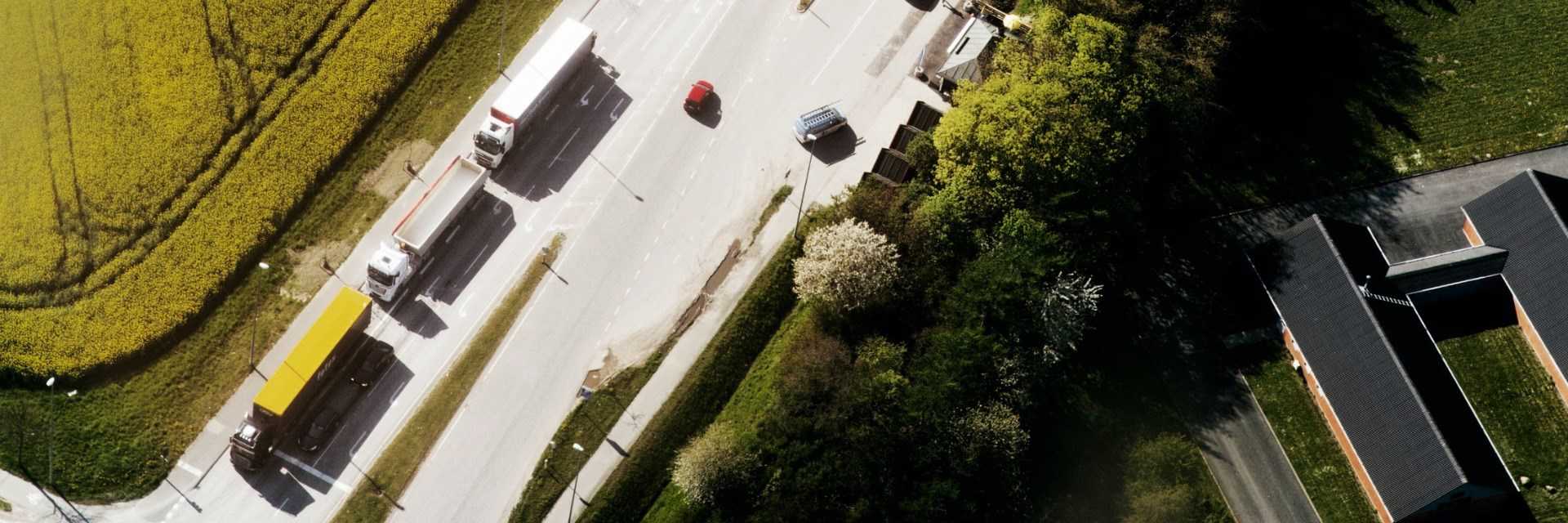 Luftfoto af en vej med tre lastbiler og fire personbiler. På begge siden af vejen er der grønne områder.