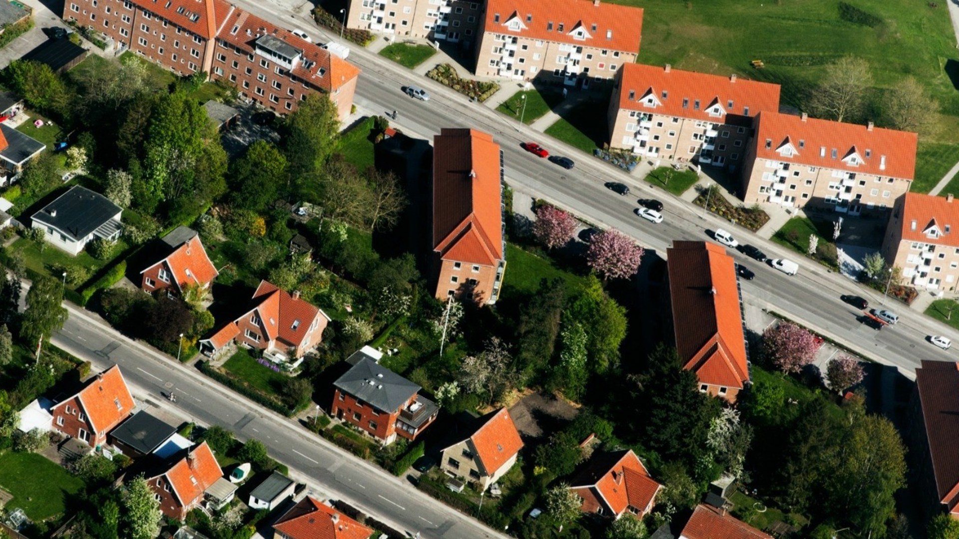 Luftfoto af boligblokke med røde tage, omgivet af grønne træer og to veje med bile.
